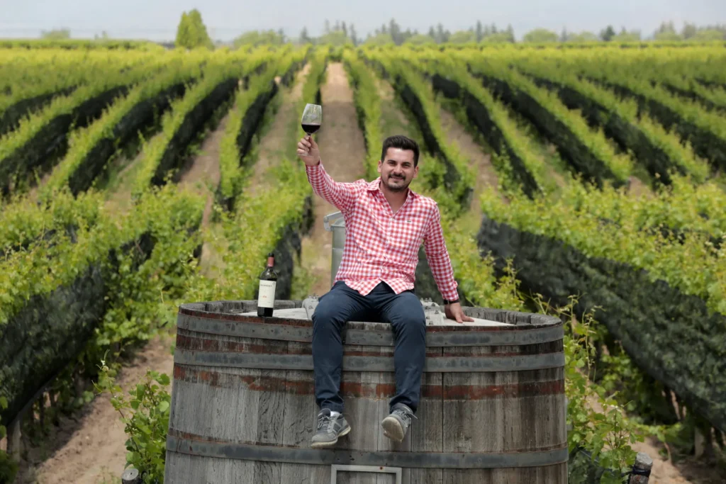 Man sitting on a wine barrel in the middle of a vineyard raising a glass of red wine for a toast