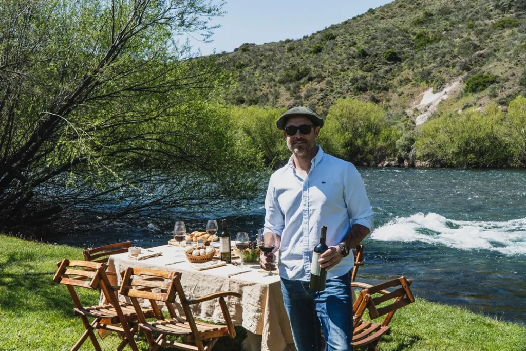 Host standing by a beautifully set riverside lunch table holding a bottle of wine